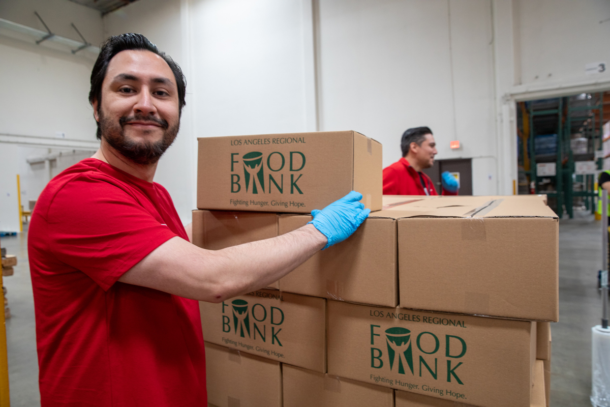 A man in a red shirt and blue gloves stands next to stacked boxes labeled "Food Bank" in a bustling Los Angeles Regional Food Bank warehouse. He is smiling at the camera, while another person is visible in the background.