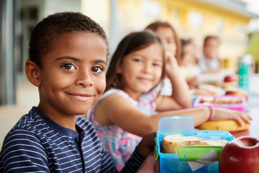 monthly-giving A group of children is sitting at a table, smiling while enjoying a meal from the Los Angeles Regional Food Bank. The focus is on a boy in the foreground with short hair and a striped shirt. Lunch boxes and food items are visible on the table, while the background remains slightly blurred.