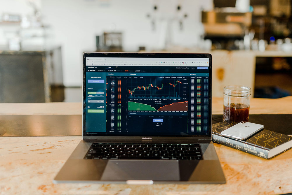 stock-lafb A laptop on a wooden table displays a financial trading platform with charts and graphs. Beside it, supporting the Los Angeles Regional Food Bank initiative, lies a branded notebook, alongside a smartphone and a glass of iced coffee. The background is a blurred workspace.