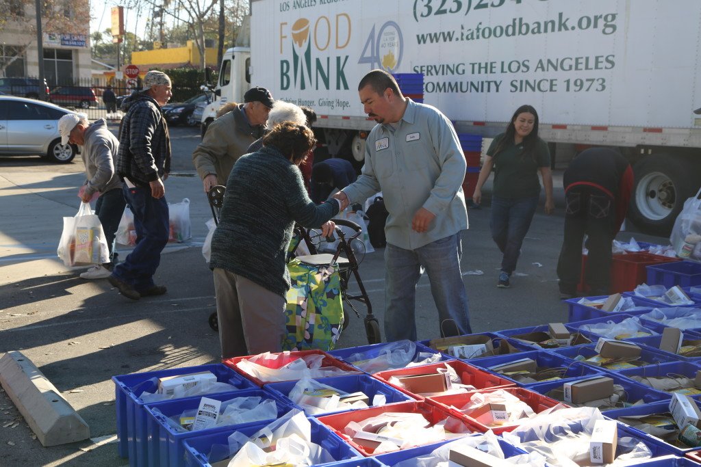 calling-all-senior-centers-we-need-your-help-la-food-bank-stories A man assists a senior woman with a walker at the Los Angeles Regional Food Bank distribution. They are surrounded by blue bins filled with food items. A truck nearby displays "Food Bank," and other volunteers are busy helping people in the background.