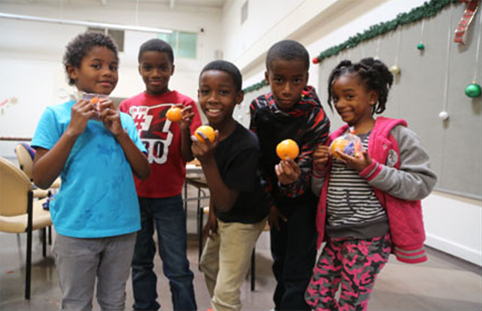 dev-fb-lar_childrens-img-v1 Five children stand indoors, smiling and holding oranges provided by the Los Angeles Regional Food Bank. They are dressed in casual, colorful clothing. The background features a decorated wall with ornaments hanging, reflecting a cheerful and generous spirit.
