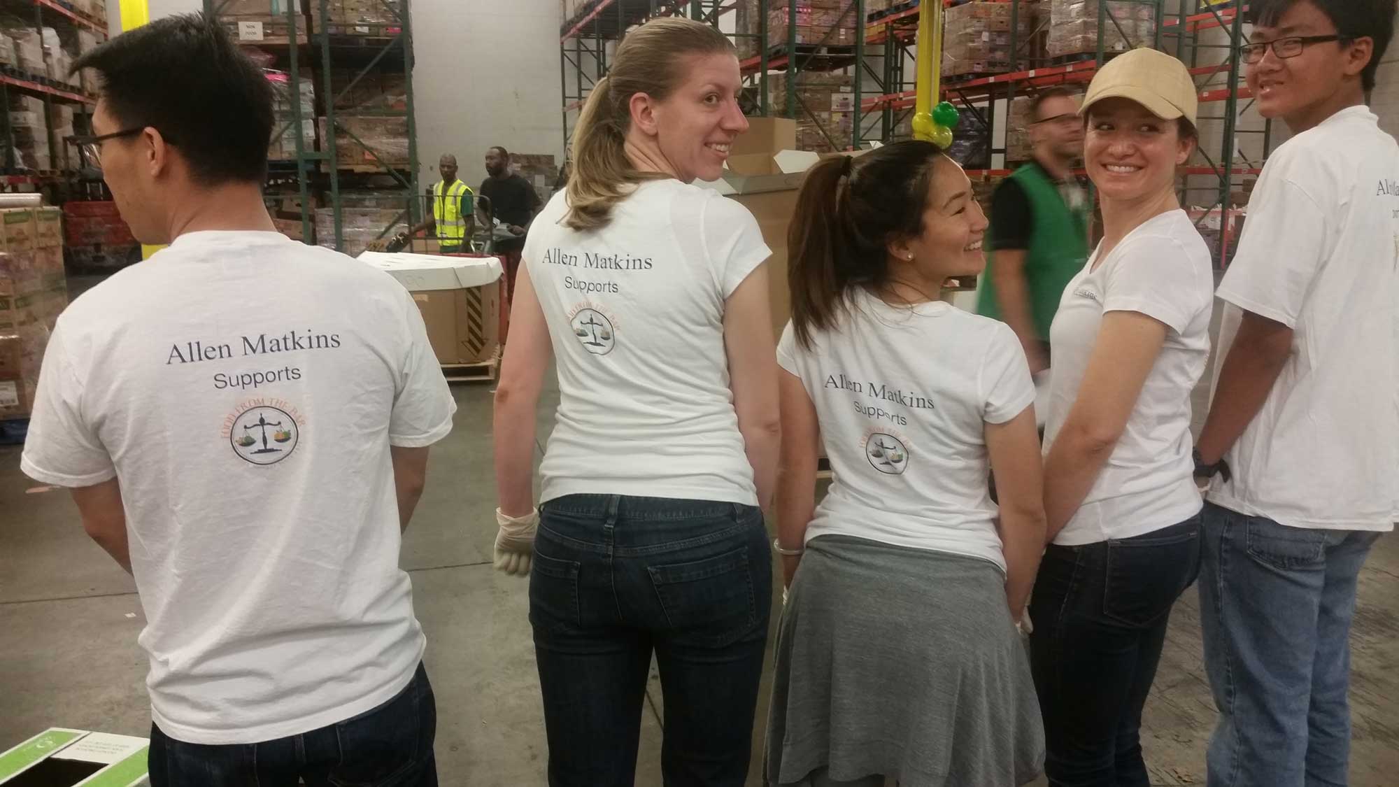FFTB Family & Friends Day 4-25-2015 (13) A group of five people wearing matching white shirts with "Allen Matkins Supports" printed on the back stand smiling in a Los Angeles Regional Food Bank warehouse, surrounded by stacks of boxes and shelves.