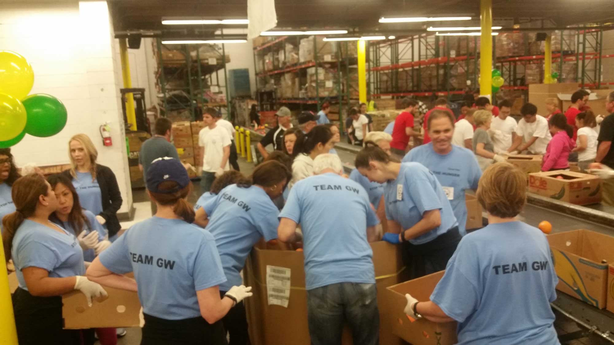 FFTB Family & Friends Day 4-25-2015 (10) A group of people in blue "TEAM GW" shirts hustle efficiently at the Los Angeles Regional Food Bank, organizing items into cardboard boxes. The industrial space buzzes with activity, rows of shelves lining the background under bright overhead lighting.