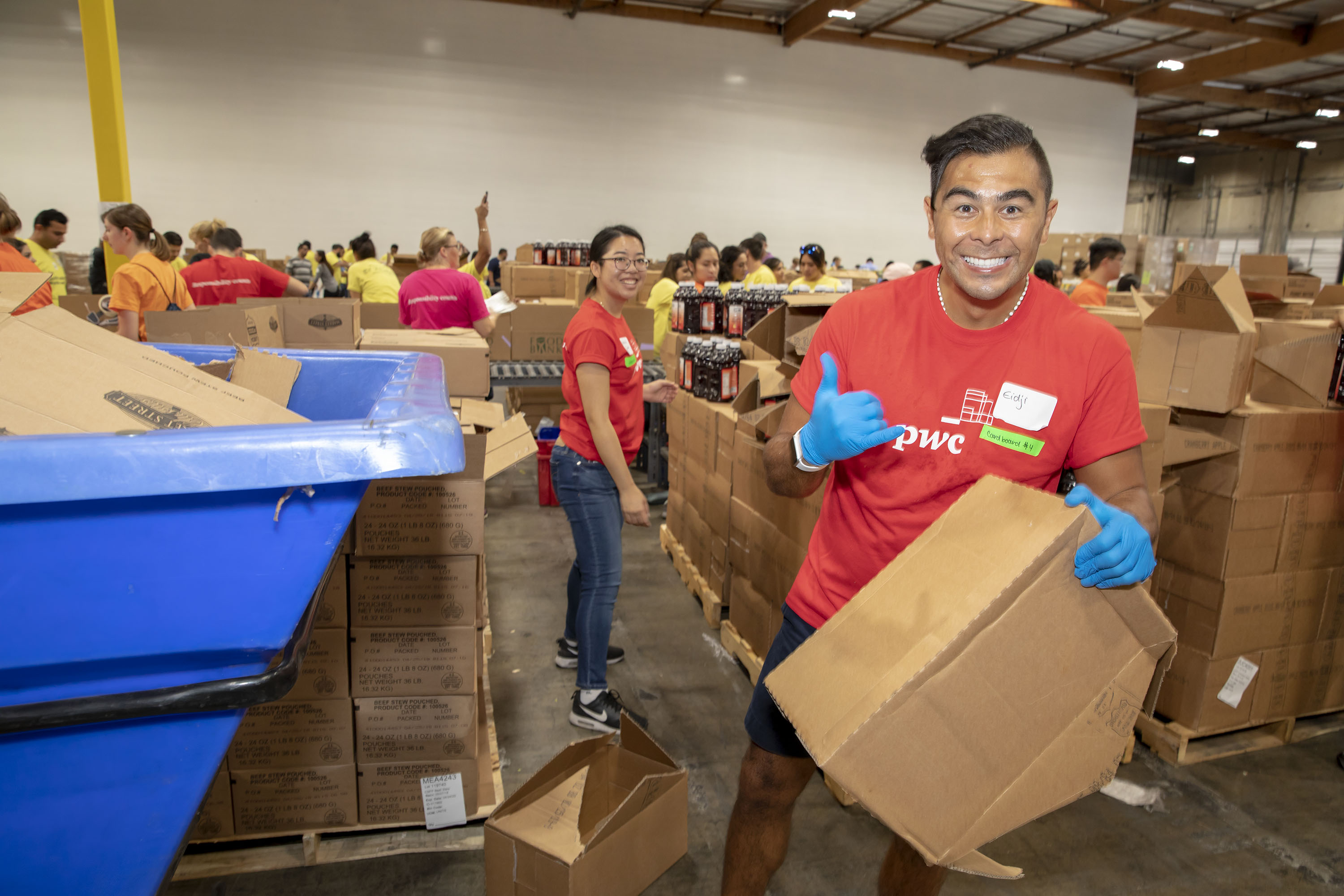 072818_BigFour2018_0999 A group of volunteers at the Los Angeles Regional Food Bank is working in a warehouse, sorting cardboard boxes. One person in the foreground is smiling and giving a thumbs-up, while others in the background are actively involved in organizing and packing.