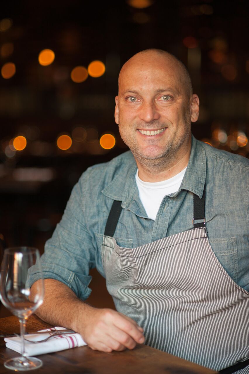 steve samson A man wearing a denim shirt and striped apron smiles while sitting at a table, reminiscent of community gatherings at the Los Angeles Regional Food Bank. A folded napkin and empty wine glass rest before him, against a softly blurred background with warm lights.