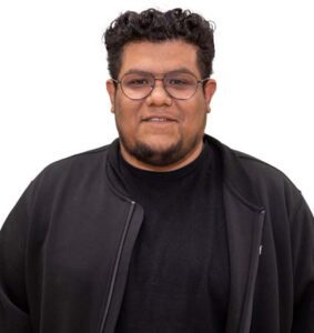 A person with curly hair and glasses, wearing a black jacket over a black shirt, is smiling warmly as they support the Los Angeles Regional Food Bank against a plain white background.