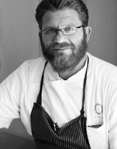A bearded man with glasses is wearing a white chef's coat and a dark striped apron, standing proudly at the Los Angeles Regional Food Bank. He smiles slightly, facing the camera in this captivating black and white photo.