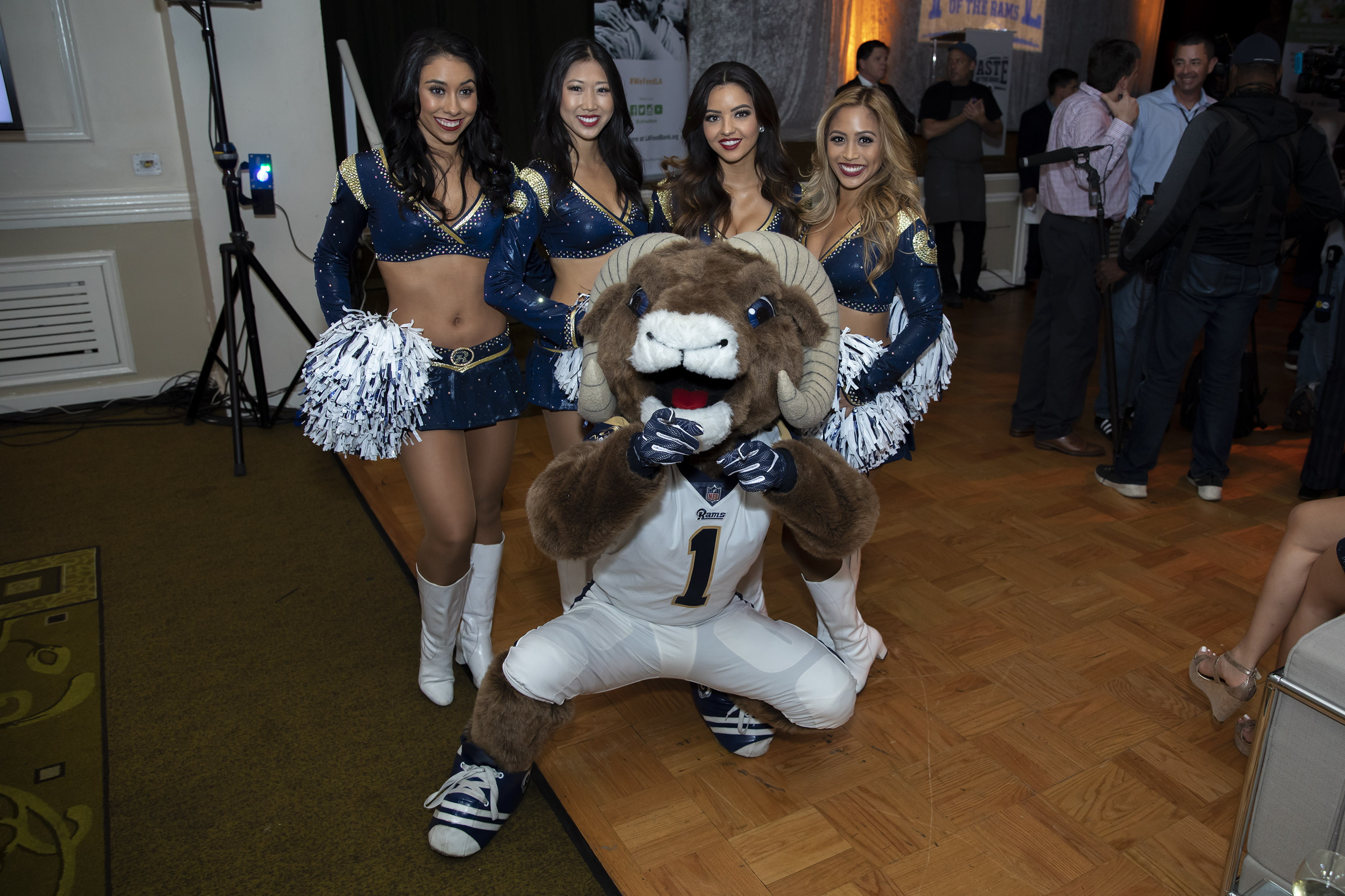 A costumed sports mascot wearing a jersey poses with four cheerleaders in blue and silver uniforms holding pom-poms. They are indoors on a wooden floor with people in the background, supporting an event for the Los Angeles Regional Food Bank.