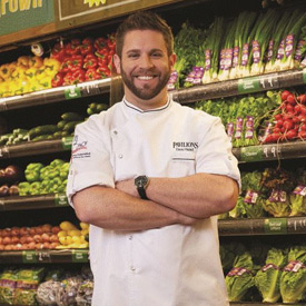 foodbank-tasteoftheRams_dave-histed_pavilions-275x275-v1 A smiling man in a chef's coat stands with arms crossed in front of a display of fresh vegetables at the grocery store, reminiscent of the abundant shelves at the Los Angeles Regional Food Bank. Lettuce, peppers, and onions burst forth with vibrant color and promise.