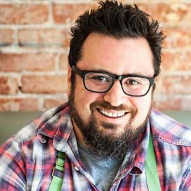 foodbank-tasteoftheRams_bruce-kalman-275x275-v1 A bearded man with glasses and a friendly smile, wearing a plaid shirt and green apron, stands against a brick wall background. He's proud to volunteer at the Los Angeles Regional Food Bank, dedicating his time to help the community.
