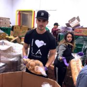 la-food-bank_my-favorite-meal-Wefeedla-4-180x180 A person wearing a black cap and "I ❤️ LA" shirt is holding a bag of potatoes at the Los Angeles Regional Food Bank. In the background, another individual manages a loaf of bread, surrounded by stacks of boxes and packaged goods.