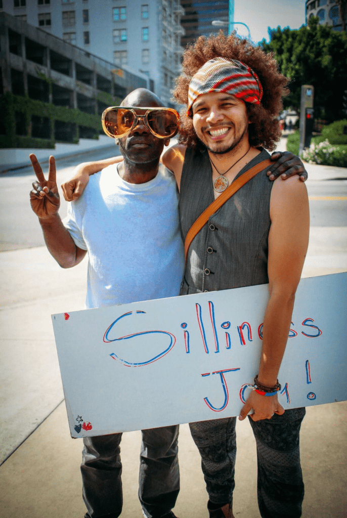 Two men smiling and posing on a bustling Los Angeles street. One sports oversized sunglasses and flashes a peace sign, while the other holds a sign reading "Silliness = Joy!" Their infectious joy echoes the spirit of giving, reminiscent of efforts at the Los Angeles Regional Food Bank.