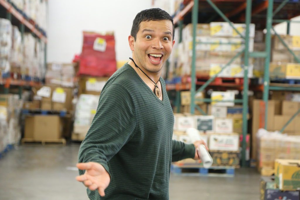 A person with a cheerful expression stands in a warehouse filled with boxes and pallets at the Los Angeles Regional Food Bank. They are wearing a green long-sleeve shirt and holding a rolled-up paper, arms slightly outstretched, ready to make an impact.