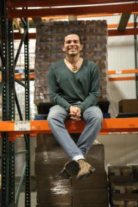 A person in a green sweater and jeans is sitting and smiling on an orange metal beam at the Los Angeles Regional Food Bank warehouse. Behind them are stacked boxes, with metal shelves and industrial lighting enhancing the scene.