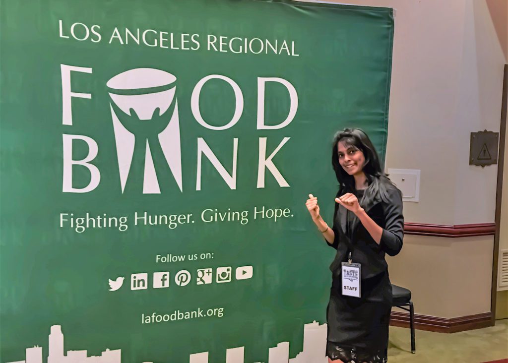 A woman stands smiling in front of a green backdrop emblazoned with the Los Angeles Regional Food Bank logo and the motto "Fighting Hunger. Giving Hope." Wearing a staff badge, she points to the logo, standing indoors with a chair beside her, embodying the spirit of community support.