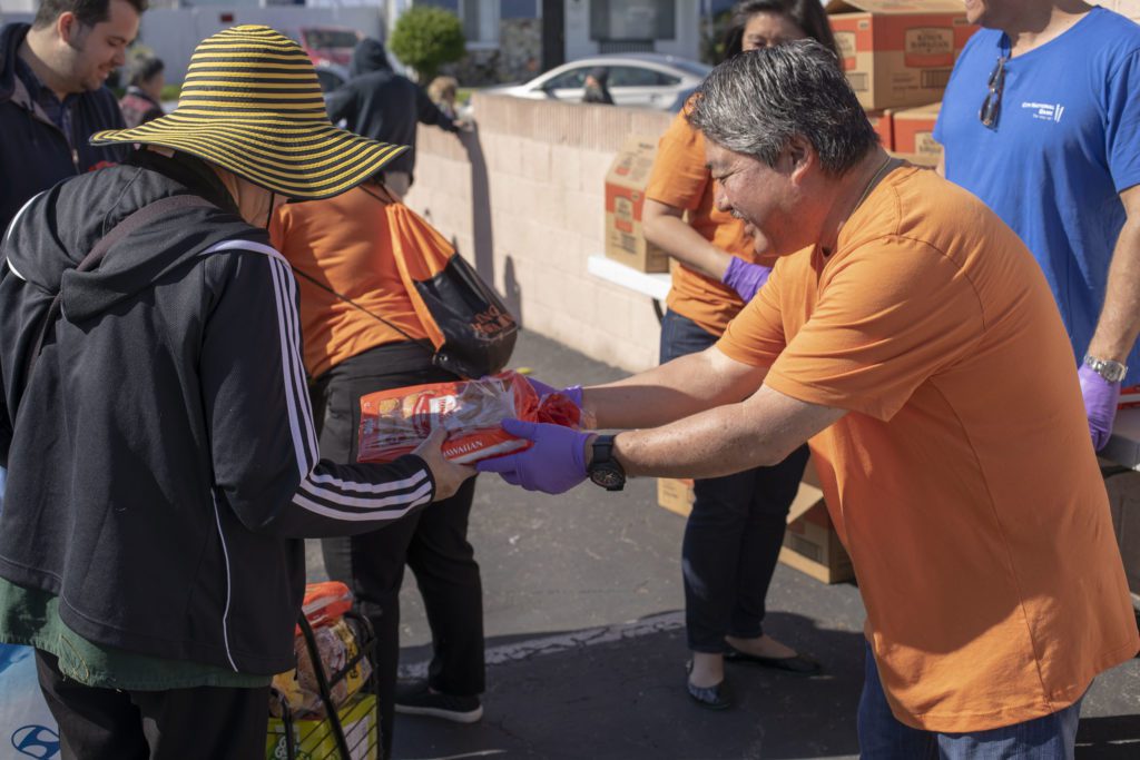 A volunteer in an orange shirt hands a bag of food to a person wearing a striped hat at an outdoor Los Angeles Regional Food Bank distribution event. Other volunteers and boxes are visible in the background.