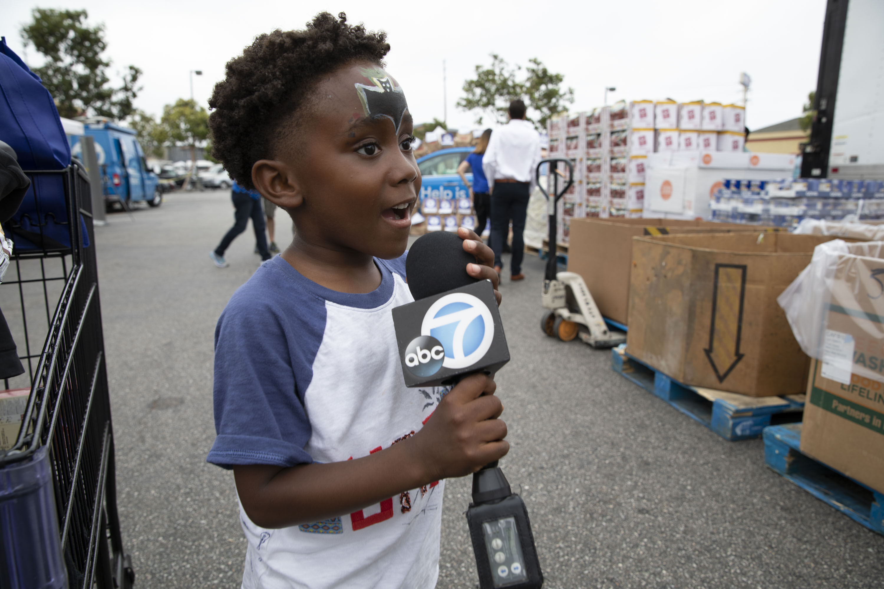 062218_FeedSoCal_9655 A young child with face paint holds a microphone labeled "ABC 7" while standing outdoors near a pile of boxes and carts. The scene buzzes with activity as people and vehicles gather at the Los Angeles Regional Food Bank event, highlighting a community coming together.