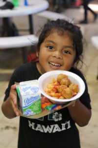 A child smiles while holding a plate of spaghetti with meatballs and a carton of low-fat milk, proudly wearing a black T-shirt with "Hawaii" printed on it. Against the backdrop of tables, this joyful scene at the Los Angeles Regional Food Bank embodies the spirit of community and nourishment.