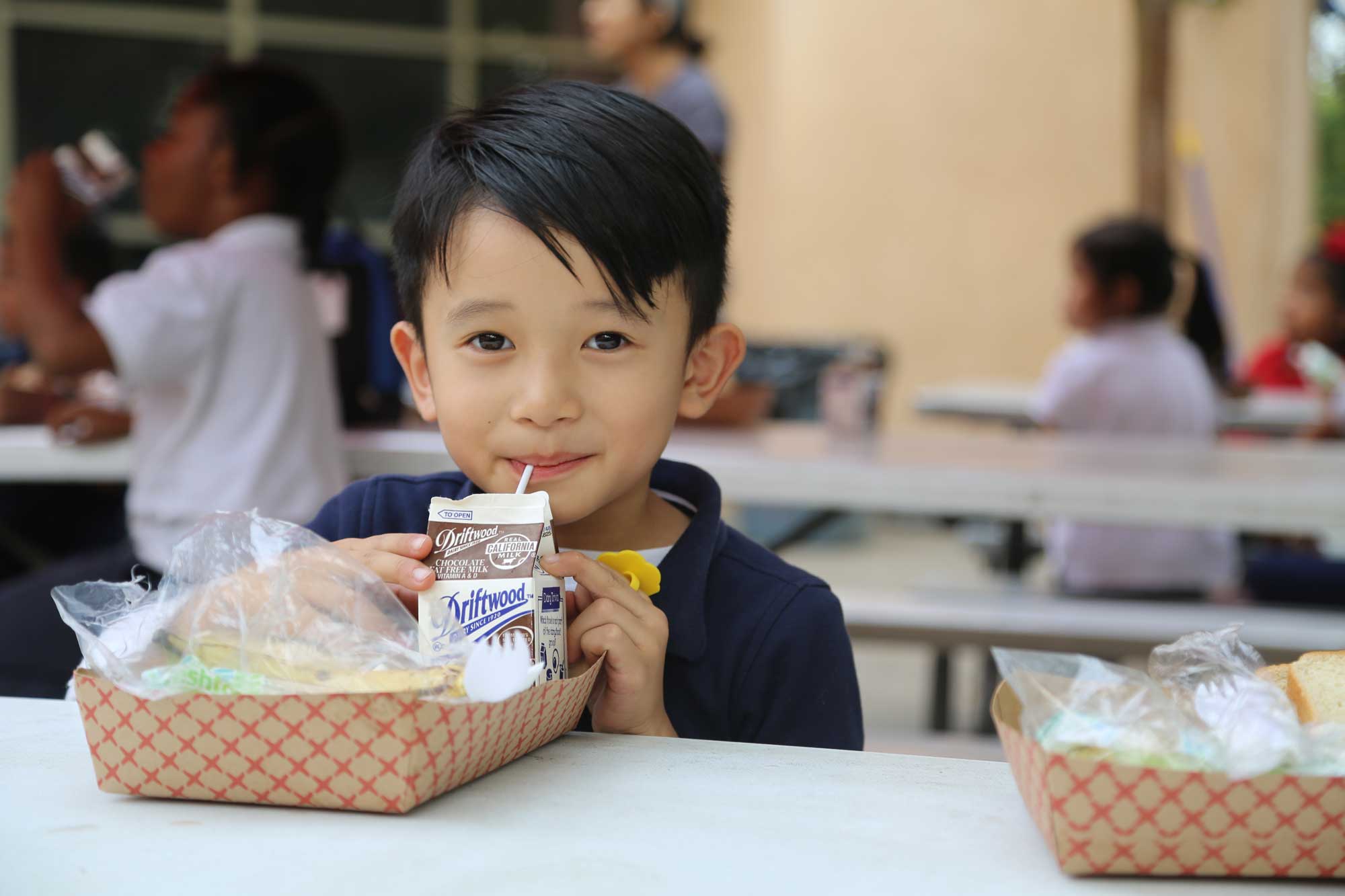 W31A3583 A young boy sits at a table with a lunch tray, eagerly sipping from a carton of chocolate milk with a straw. Around him, other children chat in the bustling school cafeteria, supported by efforts like the Los Angeles Regional Food Bank to ensure nutritious meals for all.