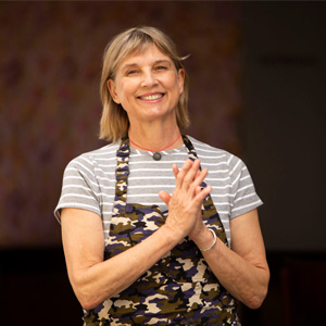 Mary_Sue_Milliken-300-300 A person smiling warmly, wearing a striped shirt and a camouflage-patterned apron, stands with hands clasped in front, embodying the spirit of the Los Angeles Regional Food Bank. The background is softly blurred, capturing their dedication and warmth.