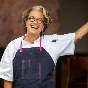 Susan-Feniger-300-300 A smiling woman with curly hair and glasses, wearing a white chef's coat and a navy apron with pink trim, holds one arm up. She stands proudly in front of the Los Angeles Regional Food Bank backdrop.