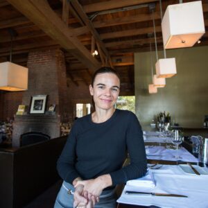 A person with dark hair is smiling and leaning on a set table in a restaurant, reminiscent of those that support the Los Angeles Regional Food Bank. The room features wooden beams, a brick fireplace, and pendant lights. The table is elegantly set with white tablecloths, plates, and glasses.