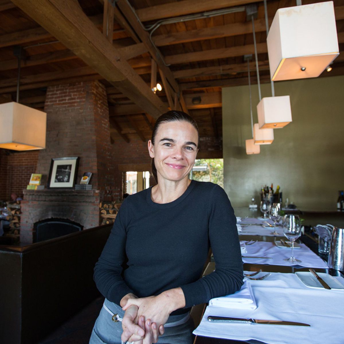 Suzanne_Goin.JPG A person with dark hair is smiling and leaning on a set table in a restaurant, reminiscent of those that support the Los Angeles Regional Food Bank. The room features wooden beams, a brick fireplace, and pendant lights. The table is elegantly set with white tablecloths, plates, and glasses.