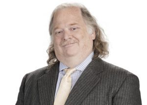 A person with shoulder-length hair and a mustache smiles at the camera, exuding confidence in a pinstripe suit with a light-colored shirt and tie. This image supports the Los Angeles Regional Food Bank's mission to fight hunger, set against a plain white backdrop.