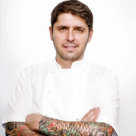 LA-foodbank-Ludo-Lefebvre-275x275-v1 A man with tattoos on his forearms stands confidently with arms crossed, clad in a white chef's jacket. His dedication, evident in every detail, reflects his commitment to the Los Angeles Regional Food Bank, against a seamless white background.