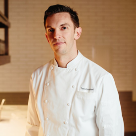 LA-foodbank-travis-strickland-275x275-v1 A chef in a white uniform stands in a warmly lit kitchen, reminiscent of the bustling atmosphere at the Los Angeles Regional Food Bank. The background features white tiles and essential kitchen equipment, with the chef's short dark hair and slight smile adding a personal touch.