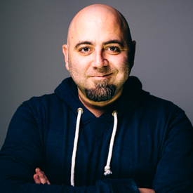 LA-foodbank-duff-goldman-275x275-v1 A bald man with a goatee, wearing a navy blue hoodie with white drawstrings, stands against a gray background. He's smiling warmly, arms crossed, embodying the spirit of community as he supports the mission of the Los Angeles Regional Food Bank.