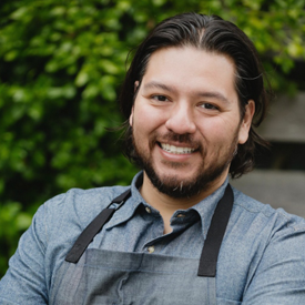 LA-foodbank-ray-garcia-275x275-v1 A smiling man with long dark hair and a beard, wearing a blue shirt and a gray apron, stands outdoors with a background of green foliage, volunteering for the Los Angeles Regional Food Bank.