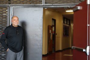 A man stands smiling near the open door of a hallway, sporting a black long-sleeve shirt with sunglasses hanging from it. The Los Angeles Regional Food Bank's emblem glints on his sleeve. The hallway features a red floor and cream walls, with a wall display visible inside.