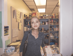 A person stands in front of a small food pantry linked to the Los Angeles Regional Food Bank, stocked with various canned and boxed items. They are smiling, wearing a gray top, and carrying a black shoulder bag. The shelves behind are neatly organized with food products.