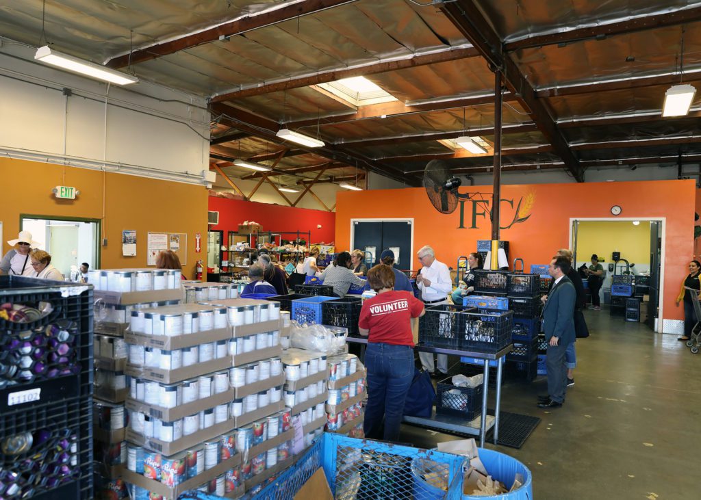 At the bustling Los Angeles Regional Food Bank, volunteers assist as people collect supplies. Stacks of canned goods and packaged foods are neatly organized in crates. The room features high ceilings and brightly painted walls, with a volunteer in a red "volunteer" shirt actively helping others.