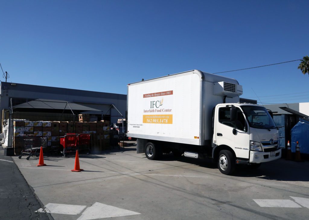 A white delivery truck from the Los Angeles Regional Food Bank is parked next to a warehouse. Boxes are stacked near the open unloading area, with orange traffic cones and shopping carts visible. The truck is marked with "IFC Interfaith Food Center" and a phone number. The sky is clear and blue.