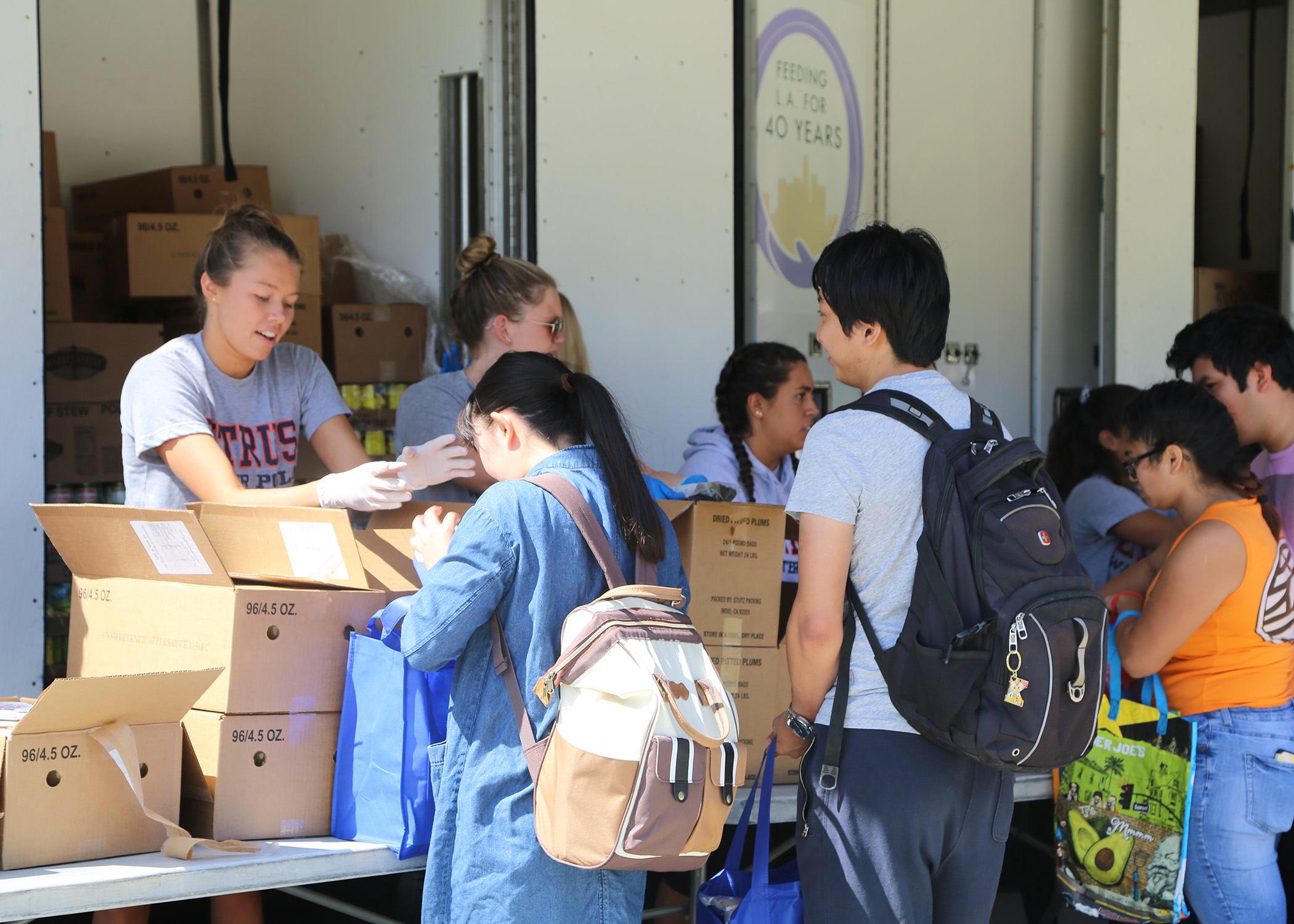 People are gathered around a table, packing boxes with supplies for the Los Angeles Regional Food Bank. Some are wearing backpacks, suggesting they are volunteering. A truck behind them has a logo with text indicating a food distribution event.
