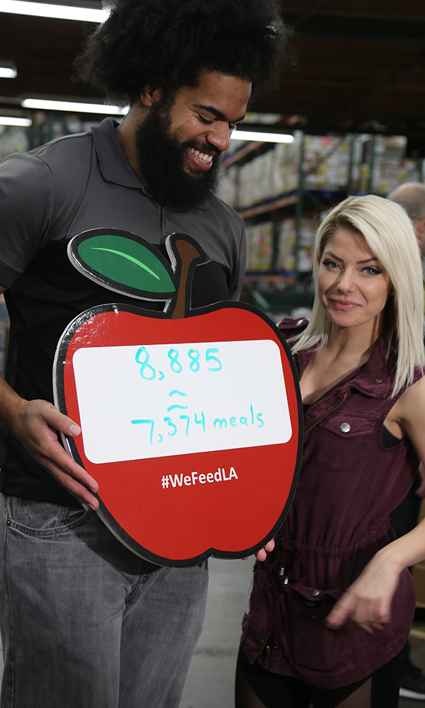 Two people hold a sign shaped like an apple reading "8,885" and "7,374 meals," with "#WeFeedLA" proudly displayed. Shelves stacked with boxes sit in the background. The woman has blonde hair, while the man's big smile shines. This initiative is a part of the Los Angeles Regional Food Bank's efforts.