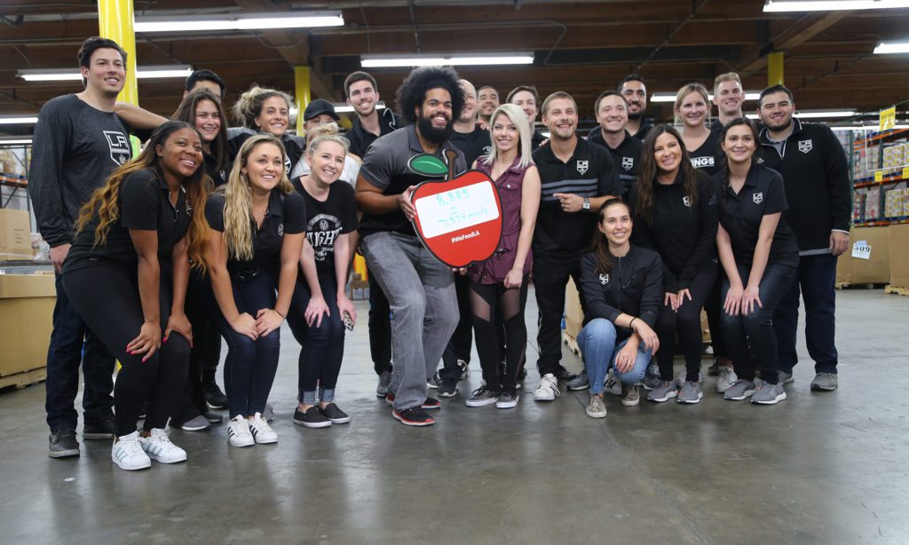 A diverse group of people wearing black shirts gathers in a warehouse, smiling and holding a large apple-shaped sign that reads "Feed the Funnel" for the Los Angeles Regional Food Bank. Shelves and boxes are visible in the background, reflecting their shared commitment to fighting hunger.