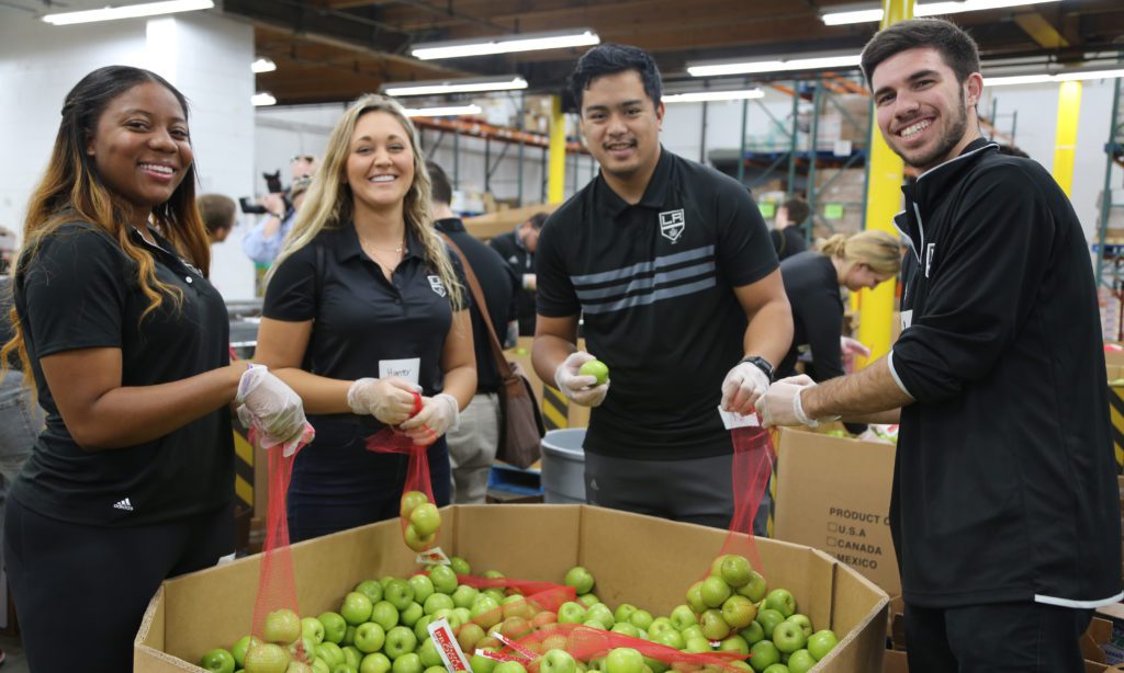 Four people smiling while packing apples into bags at a Los Angeles Regional Food Bank charity event. They stand around a large container filled with green apples, wearing black shirts and gloves. The background reveals the bustling setting of a busy warehouse.