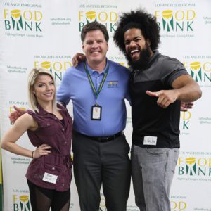 Three people smiling brightly at a Los Angeles Regional Food Bank event pose in front of a backdrop featuring the charity’s logo. Two wear name tags, while one warmly wraps an arm around another, radiating camaraderie and support for this vital community cause.