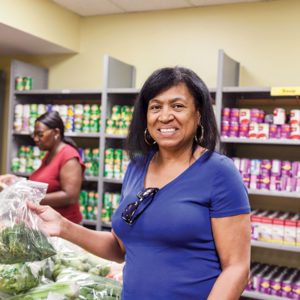 A woman smiles while holding a bag of vegetables at the Los Angeles Regional Food Bank. In the background, another person arranges items on shelves stocked with canned goods, ensuring everything is ready for those in need.