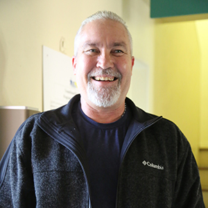 A man with short gray hair and a beard is smiling broadly. Wearing a dark fleece jacket over a navy shirt, he stands indoors against a light-colored wall with a green and yellow backdrop, embodying the spirit of community found at the Los Angeles Regional Food Bank.