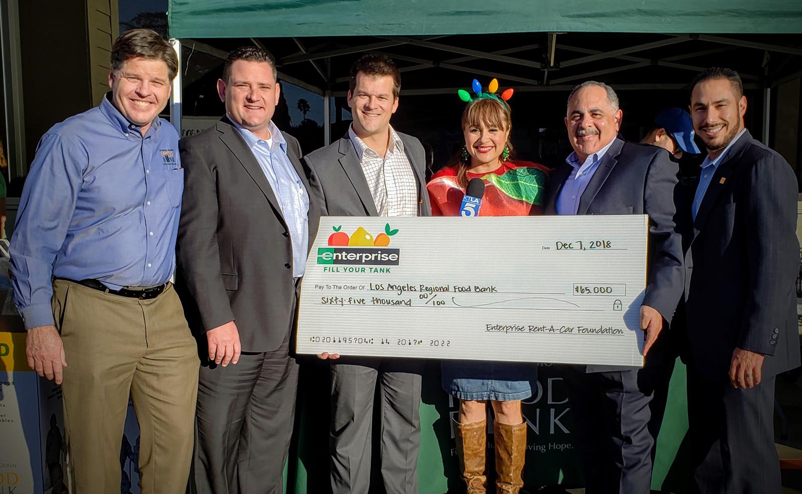 A festive group of six smiles warmly, gathered around a large ceremonial check for $45,000 addressed to the Los Angeles Regional Food Bank. Dated December 7, 2018, the scene is brightened by one member donning a cheerful red and green costume.