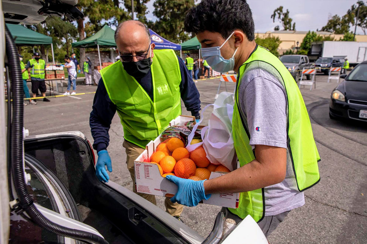 lafb-volunteer-pantry Two volunteers in bright green vests are placing a box of oranges into a car trunk at an outdoor Los Angeles Regional Food Bank event. They are both wearing masks and gloves, with several cars and tents visible in the background.