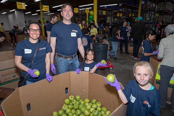 A group of people, including adults and children, are proudly wearing matching shirts and gloves while volunteering at the Los Angeles Regional Food Bank. They stand near large bins filled with green apples in a warehouse setting, some holding apples and smiling warmly at the camera.