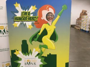 At the Los Angeles Regional Food Bank, a person stands behind a superhero cutout, their smile peeking through the face hole. Dressed in vibrant green and yellow, the superhero proudly declares, "I'M A HUNGER HERO!
