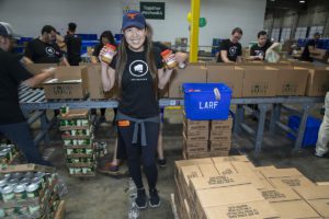 A volunteer wearing a cap smiles while holding two jars of peanut butter in a bustling warehouse. Around her, dedicated team members pack food into boxes labeled "Los Angeles Regional Food Bank." Shelves brimming with canned goods create the backdrop for this spirited scene.