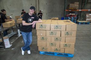 A smiling person in a cap and black T-shirt leans against a stack of boxes labeled "Food Bank" on a blue pallet in the Los Angeles Regional Food Bank warehouse. In the background, individuals are busy packing more boxes on tables.