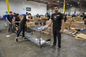 A group of volunteers efficiently packs food boxes in a bustling Los Angeles Regional Food Bank warehouse. A man stands beside a table filled with supplies, as the background showcases stacked boxes and a prominent "Food Bank" sign, reflecting an environment that is both busy and organized.